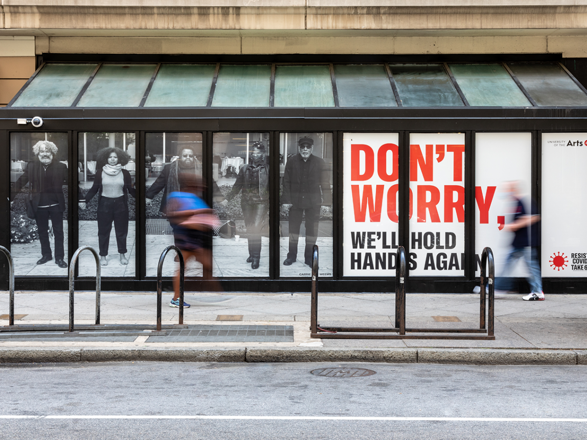 一块广告牌上，人们手牵着手，用红色字体写着“别担心，我们还会再牵手的”。