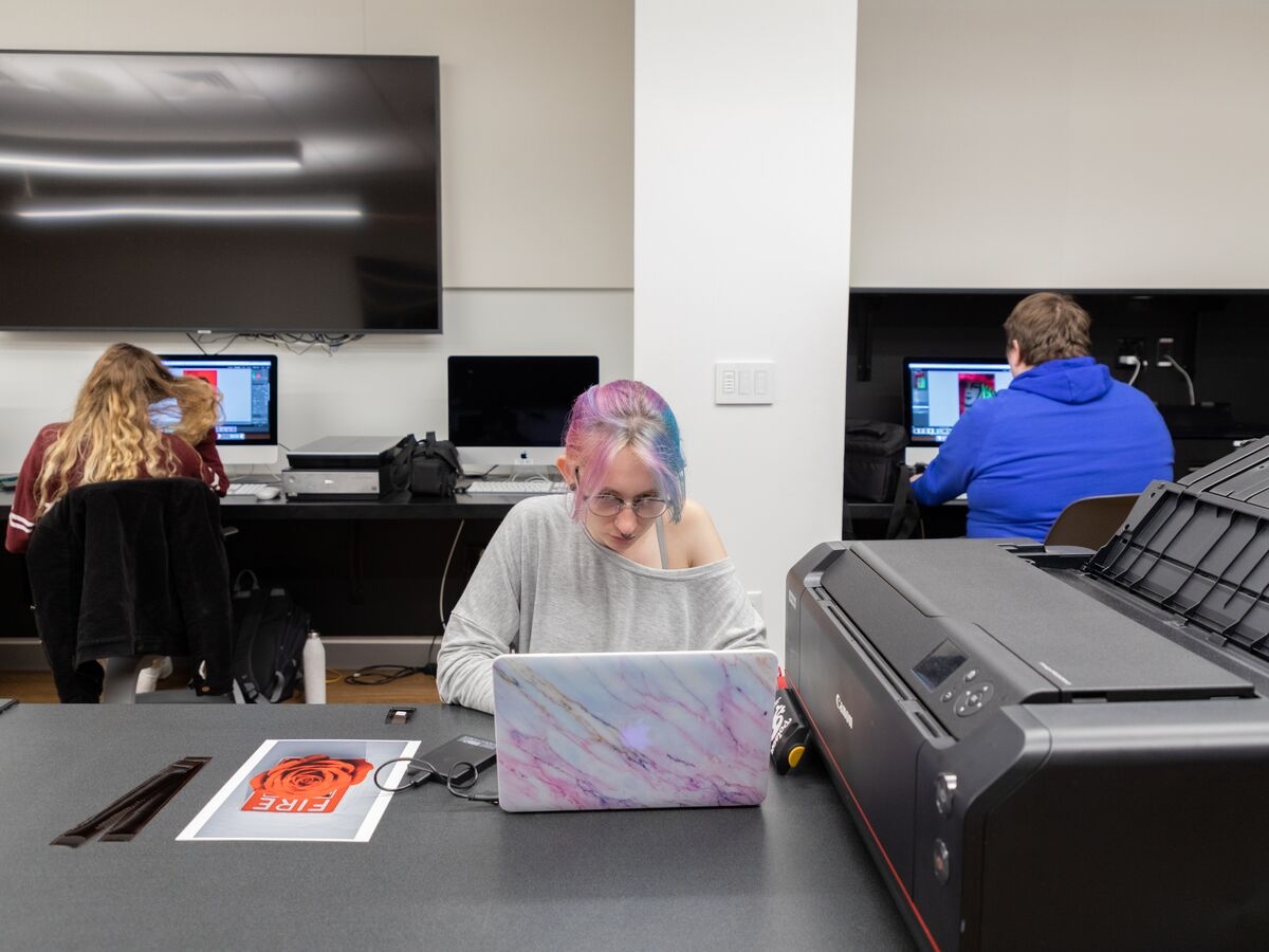 在数字成像实验室里，一个学生在打印机旁的笔记本电脑上工作。