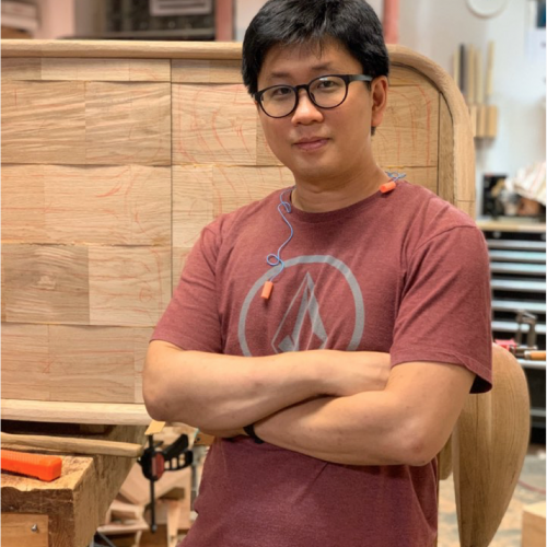 Keunho Peter Park穿着一件玫瑰色的t恤，双臂交叉站在一件未完成的家具前。