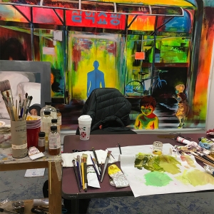 学生的绘画工作室。颜料、刷子、纸巾和其他用品横放在桌子对面，后面的墙上挂着一块画布。