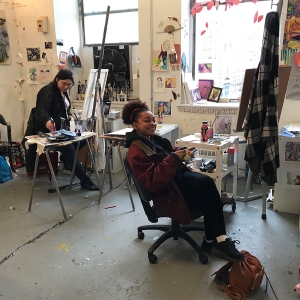 两个学生在画室里画画。一个学生一边工作一边对着镜头微笑。