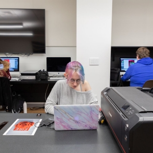 在数字成像实验室里，一个学生在打印机旁的笔记本电脑上工作。